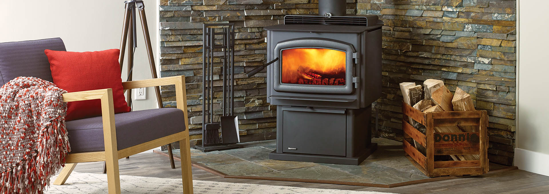 Regency F2500 Wood Stove lit in front of staggered stone heat shield with living room chair and wood pile next to it